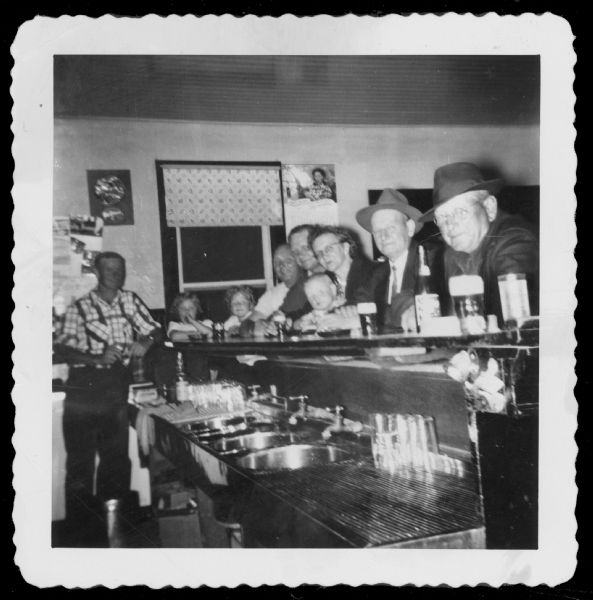 View from behind bar towards a man standing on the left, and a group of men, women and children sitting at the bar.

Pictured from left to right are Arnie Wittnebel (Roy's brother), Joyce and Barb (daughters of Roy), Roy Wittnebel, Lorenz Wittnebel (cousin), Deloris Wittnebel, Tom Wittnebel (son of Roy), Herman Generman (friend /customer), and Frank Wittnebel.