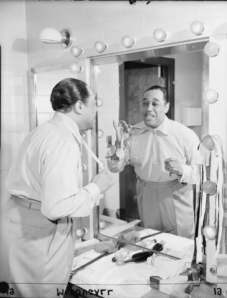 Duke Ellington is knotting his necktie in a mirror prior to a performance at the Milwaukee Arena.