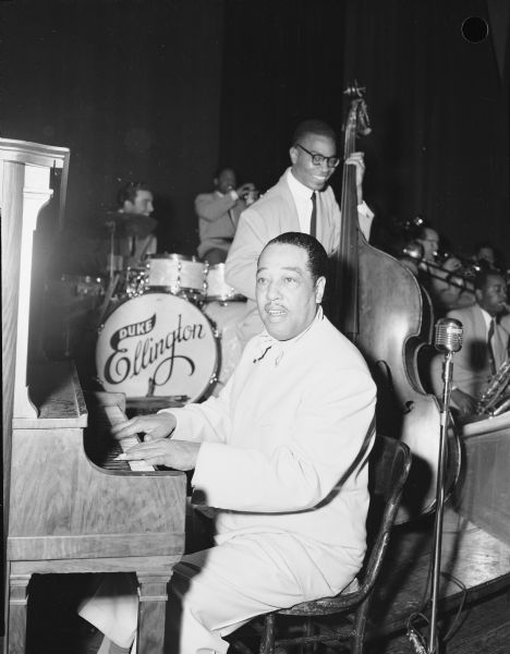 Duke Ellington at the piano during a performance with the Duke Ellington Orchestra. The bassist is likely Joe Benjamin, and the drummer is Louie Bellson. Other orchestra members are in the background.