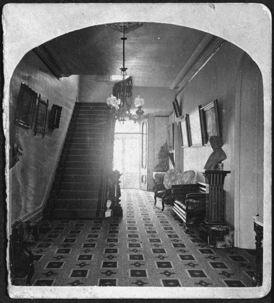 One half of a stereoview of the first floor hallway of Villa Louis. The hallway contains a bust of Hercules Dousman, a chair, a couch, and several paintings. A light fixture is hanging from the ceiling near the base of the stairs.