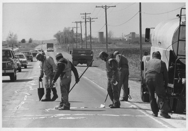 Five men are working on a highway, placing tar and covering it with tissue. A line of cars are passing in the opposite lane. A field and farm buildings are in the background. Caption reads: "SPRING-TIME PROJECT — Washington County Highway Department workmen spread tar over cracks on Highway 167, west of Highway 145 in Germantown Wednesday. The workers cover the tar with bathroom tissue, in order to protect vehicles from being splashed by the tar."