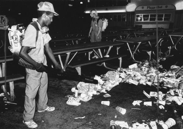 A man with a backpack-powered leaf blower is blowing trash along the ground between picnic tables. Another man with a similar blower is in the background. Further back, a food stand has the Pizza Hut logo. Caption reads: "Larry Johnson blew trash out from under tables during the night shift at Summerfest."