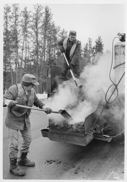 Two men are shoveling steaming asphalt from the rear of a truck. Caption reads: "Portage County Highway Department workers Victor Peters (left) and Arleigh Schroeder mixed a steaming batch of new asphalt as they prepared to repair pot holes caused by winter freezing on Highway 66, east of Stevens Point."