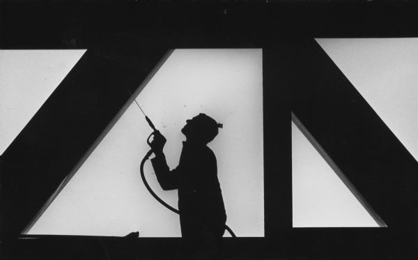 Silhouetted profile of a man looking up and spraying part of a structure with vertical and diagonal support beams with a spray gun. Caption reads: "Tim Johnson of Ace Window Cleaning sprayed the First Wisconsin Building overpass above Michigan Ave. They were cleaning the structure."