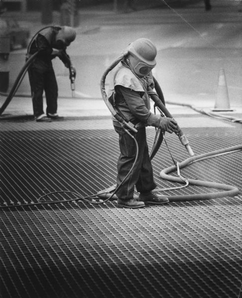 Two men in protective suits are using hoses to spray paint onto a bridge grating. Caption reads: "<b>NEW PAINT, SAME COLOR</b> — The State St. bridge over the Milwaukee River is receiving a new coat of green paint from workers, who include Ted Thompson (foreground) and Bob Bernel. The paint job is expected to take most of the summer."