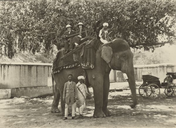 Five people are posing around an elephant in front of a large tree. Two people are standing next to the elephant, one man is the mahout sitting at the elephant's neck, and two people are sitting in a howdah (carriage). Two wheeled carriages or rickshaws are in the background on the right. Caption reads: "F. Dorr Bradley, Eleanor Bradley, Guide, Servant, Elephant Rider & Elephant, all on their way to Florence Italy to spend Easter. Jaipur, India, February 20th, 1923."