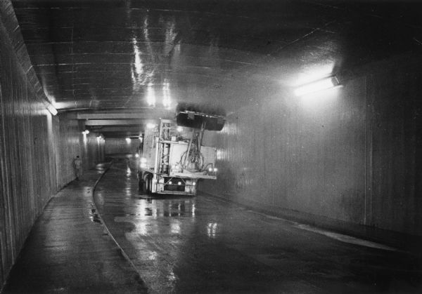 A truck is moving along a tunnel, with a rotating scrub brush attached to its rear scrubbing the ceiling of the tunnel. A man is walking on the opposite side of the street. Caption reads: "A special machine was used to scrub the interior of the tunnel taking northbound traffic from the North-South Freeway onto W. Kilbourn Ave. Both of the freeway ramps leading to tunnels running underneath MacArthur Square were closed to traffic for part of a day this week because of maintenance work."