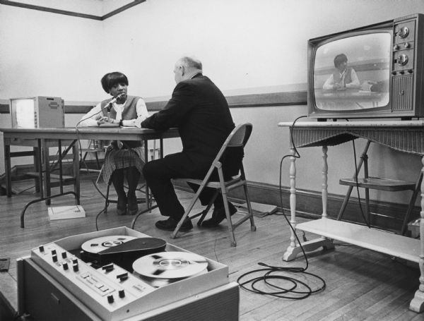 A woman and a man are sitting at a table. The woman is speaking into a microphone. A tape deck in the foreground is connected to a television. Caption reads: "Carrie Russ, 18, of 1520 N. 8th st., spoke into a microphone to record a simulated job interview with William Lepthien, employment manager for the Wisconsin Telephone Co. Senior speech pupils at Lincoln high school listened and watched her appearance shown on closed circuit television."