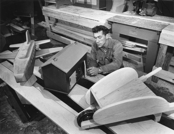 A man is sitting while examining a small wooden barn. A wooden wheelbarrow and small airplane model are adjacent to the barn. Caption reads: "A small barn, one of the products of the People's Craft Co-Operative, was checked by Gary Olson, a former migrant worker who is employed at the workshop. Olson lives at 3520 S. 92nd st."