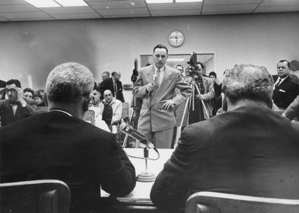 A man is standing and holding a microphone. Behind him are two cameramen with cameras, and several people sitting or standing. In the foreground are two men sitting at a table, with their backs to the camera. A microphone is between them. Caption reads: "Kenneth Roth, 34, of 2726 W. McKinley blvd., a laid off assembler at Allis-Chalmers Manufacturing Co., told [Secretary of Labor Arthur J.] Goldberg and Gov. [Gaylord] Nelson (right) that other companies would not hire him because they feared he would return to Allis-Chalmers after the recession."