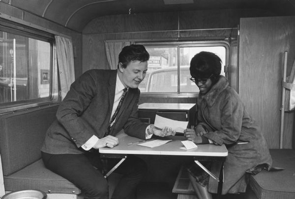 A woman and a man are sitting on either side of a table, both looking at a slip of paper the man is holding. They appear to be inside a large vehicle. Caption reads: "Mrs. Hattie Mims, 2325 N. 5th st., visited the jobmobile, where she obtained employment information from Kenneth M. Scherrer, a manpower specialist with the Wisconsin state employment service."