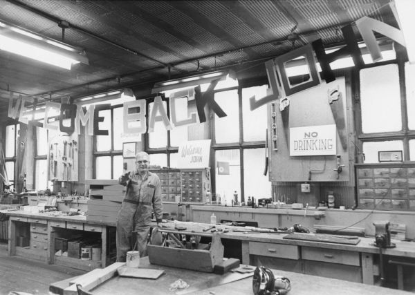 A man in work overalls is posing standing along a carpentry table, under a banner that reads: "WELCOME BACK JOHN". Additional signs read: "Welcome JOHN" and "NO DRINKING". Caption reads: "<b>RADISH BACK AT WORK</b> — Chief carpenter John Radish was greeted by banners and signs upon his return to work in mid-December. Radish, who had been ill a month with heart trouble, is 'feeling fine' now. The large letters were cut out by Jacqueline Gaulke, building department secretary."