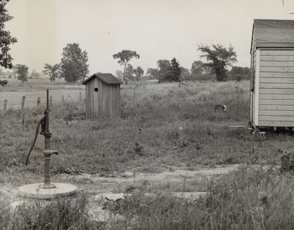 Two outhouses and another building are in a field, with a water pump in the left foreground. Caption reads: "This Press picture shows the water supply and plumbing facilities — or lack of them — at the Mexican labor settlement east of Plymouth on Highway 23. The pump and larger outhouse are easily distinguished, but you will have to look carefully into the background of the picture to find the second outhouse which is of smaller proportions."