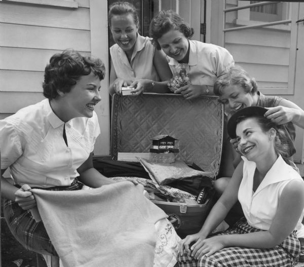 Five young women are smiling and posing around an open suitcase, which contains various items. One woman is setting a hat on another woman's head. Caption reads: "Back from a six week tour of Europe, these Wauwatosa teenagers showed their souvenirs Thursday. From left are Judith Anderson, 17, of 2244 N. 68th st.; Diane Schnarsky, 17, of 2209 N. 60th st.; Karen Christianson, 18, of 2225 N. 62nd st.; Mona Reider, 18, of 6422 Milw. av.; and Lucia Huth (trying on a beret), 16, of 2202 N. 72 st. They saved money four years for the trip and had some left at their return."