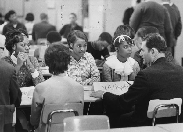 Three young women are sitting on one side of a table facing two men and a woman, whose backs are to the camera. Similar arrangements of groups are in the background. Caption reads: "Pupils at North Division high school listened as H.C. Winnick, personnel director of the veterans administration hospital (right) described job opportunities at that institution. Representatives from 24 area companies and agencies participated in job interviews at the school with 150 members of the graduating class."