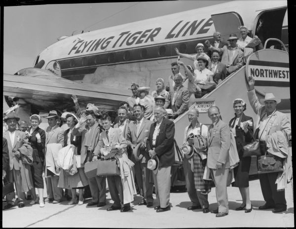 Several people posing outside a Flying Tiger Line airplane, on the ground and on the airstair. The airstair has logos for Northwest Airlines. Caption reads: "<b>The first of three planeloads</b> of Milwaukeeans left Gen. Mitchell field Friday for the national German song festival held every 10 years at Stuttgart, Germany. Altogether, 220 men and women will make the trip. There were 68 persons in the first group. A dozen German singing societies have members in two choruses which will represent Milwaukee at the event starting Thursday. Some 100,000 singers are expected at Stuttgart."