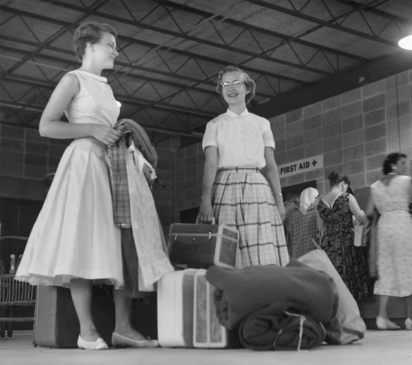 Two young women are standing in a building with their luggage at their feet. There are several women are in the background near a First Aid sign. Caption reads: "<b>Young fairgoers checked in</b> at the new youth building Saturday. They are Laura Fincher (left), 17, of Oregon, Wis., and Jo Ann Wackett, 17, of Sheboygan Falls, Wis."