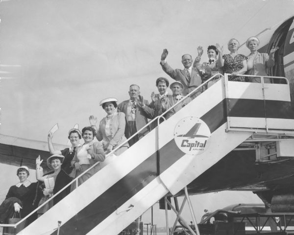 Ten women and two men posing on the airstair to a plane. Several of the people are waving. The airstair has a Capital Airlines logo. Caption reads: "<b>The first organized group</b> of Wisconsin residents to visit Poland since before World War II boarded a plane Sunday at Gen. Mitchell field for New York city. They left New York Monday on a KLM (Dutch) air liner for Poznan, Poland. From top of the ramp are Mrs. Pauline Stasielczuk, Racine; Mrs. Sophie Stabicz, 1429 W. Burnham st.; Mrs. Anton Gasiorkiewicz, 2808 N. Pierce st.; Anton Gasiorkiewicz; Mrs. Martha Philipkowski, 4304 W. Cleveland av.; Miss Josephine Philipkowski, same address; Waclaw Pokrzywa, 4736 W. Forest Home av.; Mrs. Gladys Maretto, 3777 E. Munkwitz av., Cudahy; Mrs. Laura DeBlaise, Racine; Mrs. Harriet Gostomski, 314 E. Bradley av.; Miss Marilyn Gostomski, same address, and Miss Laura Pilarski, 942 N. Cass st., a Milwaukee Journal reporter. Six other persons left earlier and will join the group in Amsterdam, Holland. They will return July 19. Almost all will visit relatives in Poland."