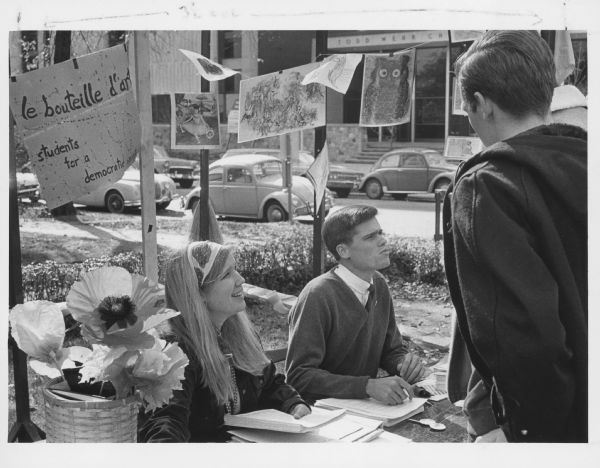 A young man and woman are seated at a booth and speaking to one or more people who have their backs to the camera. Behind them is a sign that reads: "le bouteille d'art [the art bottle], students for a democratic society." Several art pieces are suspended from string in the booth. Caption reads: "A French street scene provided atmosphere for a fair at Marquette university this week designed to acquaint faculty and students with activities. Miss Patricia Hamilton, a political science sophomore of 729 N. 11th st., and Ronald Webb, a history junior of 1310 W. Wells st., talked to visitors at the booth which was set up by the Students of a Democratic Society [sic]."