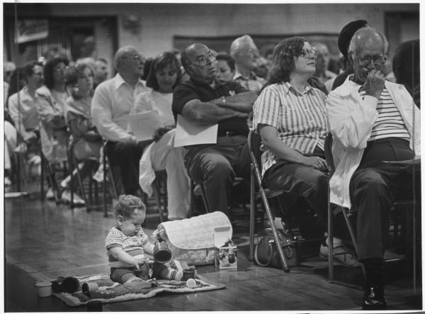 Many people are sitting on folding chairs. An infant is sitting on a blanket on the floor next to them and playing with stacking cups. Caption reads: "TOYING WITH THE IDEA — Edward Crise, 10 months, kept himself busy as his mother, Jane (right), joined about 400 people Thursday at a meeting on a municipal water system in Bayside."