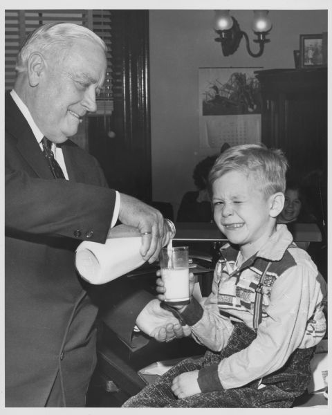 A man is pouring milk from a bottle into a glass, which is held by a boy who is winking at the camera. Caption reads: "Washington, D.C. -- Senator Alexander Wiley (R., Wis.) pours an 'extra' glass of milk for the School Milk Program. Senator Wiley is pictured here with six-year old Robert Millegan just before he left his office for the Senate Floor. Wiley urged prompt provisions of an additional $10 million of school milk funds to keep the program rolling this year. He also introduced a bill to broaden the program from the present fifty million dollars to at least seventy-five million dollars for next year."