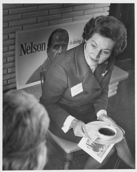 A woman is sitting and holding a cup of coffee on a plate, facing an unidentified person in the foreground. She is wearing a name tag that reads: "Carrie Lee Nelson", and on her left jacket lapel is a decorative pin, and another pin that reads: "Gaylord". Behind her is a campaign poster for Gaylord Nelson. Caption reads: "Mrs. Carrie Lee Nelson: 'I'm doing my campaign bit for Gaylord'".