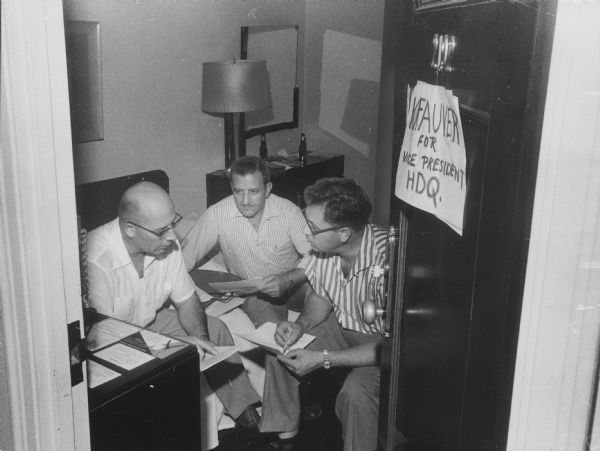 Three men are sitting in a room holding papers. A handmade sign on the door reads: "KEFAUVER FOR VICE PRESIDENT HDQ." Caption reads: "<b>A makeshift sign</b> on the hotel room door, these Wisconsin Democratic  delegates planned 'Kefauver for vice president' strategy well into Sunday night. From left are Assemblyman Howard F. Pellant, Milwaukee; James Arena, Racine, and Sam Rizzo, Racine. They held their huddle in the Blackstone hotel, where the delegates stay."