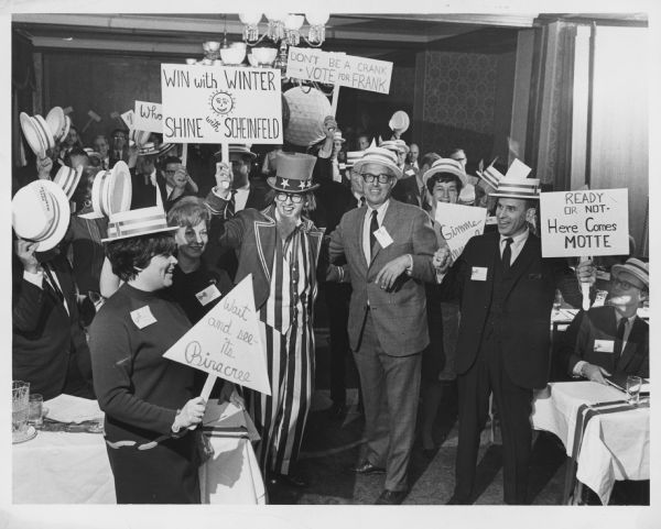 Several people wearing straw boaters are holding signs and posing with a man dressed as Uncle Sam. Signs include such slogans as: "WIN with WINTER, SHINE with SCHEINFELD", "DON'T BE A CRANK - VOTE FOR FRANK", READY OR NOT - Here Comes MOTTE", and "Wait and see - its Biracree". Caption reads: "A Manpower, Inc. sales promotion meeting had all of the hoopla and most of the trappings of a political convention here Wednesday as branch managers and franchise operators from 11 midwestern states staged a red, white and blue rally at the Pfister hotel. Uncle Sam, portrayed by Ronald Franzmeier, of the temporary help and business service firm's public relations department, and Elmer W. Winter, Manpower president, led one demonstration designed to step up sales to the fever pitch of a political campaign."