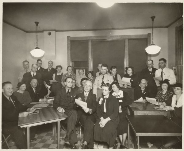 Two men and a woman are sitting in the center, with the man in the middle holding up a piece of paper that they are looking at. A large group of people are sitting or standing around them. Caption reads: "Precinct committeemen from various wards of Green Bay, Wis. From L to R - (seated) Mike Kresky, Roy Empey, Mrs. Mike Kresky. [Rest not identified]".