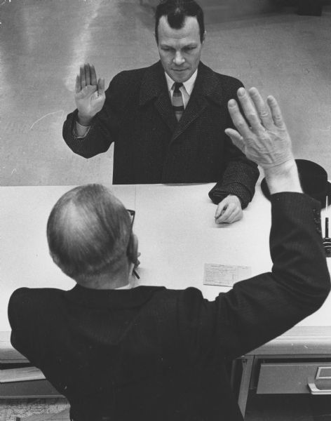 Overhead view of two men, who are standing on opposite sides of a table. They are facing each other and each has his right hand raised. Caption reads: "<b>The last registrant</b> for the primary election in Wauwatosa was Robert C. Brassard of (9533 Harding blvd.) He was sworn in by Paul H. Riemer (foreground), city clerk."