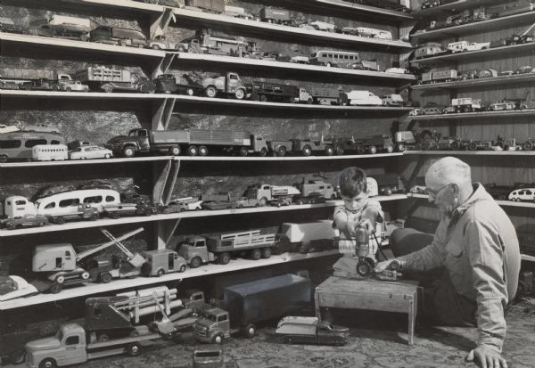 A boy is using an electric drill on part of a toy car, which is held by a man sitting beside him. Behind them are shelves full of toy cars and trucks. Caption reads: "<b>Every day</b> is paintup, fixup day for Gordon Gunderson, 8, of 1321-A W. State st. Gordon specializes in repairing old toys just for the fun of it. He has a collection now of more than 400 toys in a basement room that looks like Santa Claus' workshop. When necessary, Gordon's father, Omey, helps by shaping metal. They worked against a background of toys that would make most children bug eyed. Gordon gets the toys from rummage sales and neighbors. He said he plans to give them all away in a few years. Now, he still enjoys playing with them."