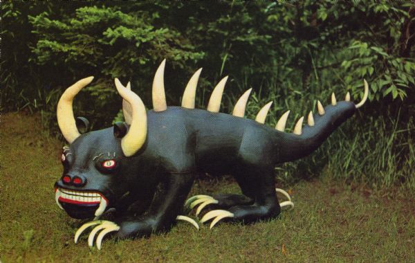 A sculpture of the mythical hodag is posed in a clearing. Caption reads: 
"RHINELANDER WISCONSIN 
'HOME OF THE HODAG'
On the first night of the arrival of lumber camp novices, an Indian from the camp would go out in a nearby swamp and make the most blood-curdling and terrifying noises. The tenderfeet were told that this was the 'Hodag' wandering in his native wilds. Many a tale was spun by the loggers about this mythical animal and a great number were taken in by the hoax."