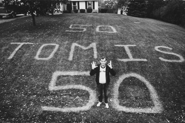 Overhead view of a man standing on a lawn. He is holding up 5 fingers on one hand and making a zero with the other. On his lawn someone has painted "50 TOM IS 50". Caption reads: "Thomas C. McFadzen stood on his surprise birthday card friends painted in his front yard."