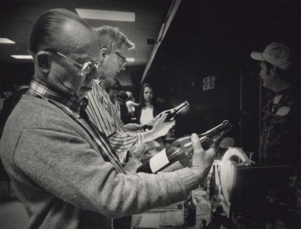 Two men examining bottles they are holding. Other people are standing in the background. Caption reads: "VINTAGE FINDS? — John Brandt (foreground) of Hartford and Dick Schwab looked over antique bottles that were on display recently at bottle and advertising sign show at the Circle B Recreation Center in Cedarburg. The bottle Brandt was holding was about 100 years old and still contained Allouez Mineral Water."