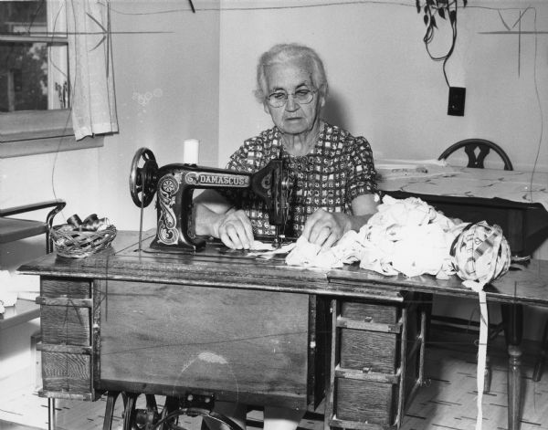 A woman is using a sewing machine on a collection of scrap fabric. Caption reads: 
"FROM RAGS TO RUGS
Our generation of grandparents make advancing age an opportunity for doing a lot of things they never had the time for previously.
Mrs. Charles [Margaret Elizabet Mulheron] Fedderly of Elmwood braids rags which she finds 'a good substitute for knitting.'
She finds that the most interesting aspect of this hobby is the many places the rags come from. A gift of drapes from St. Joseph's hospital, St. Paul, started Mrs. Fedderly on her new occupation about two years ago. Drapes from a California restaurant, sent by a daughter, went into other rugs. Another daughter in Illinois sent 14 cafe uniforms. Mrs. Fedderly also receives material from friends and neighbors and is 'always waiting to see what the next bunch will look like.'
Mrs. Fedderly is 'busy every minute,' a statement which is understandable when she explains that there is close to 400 yards of braid in an average carpet."