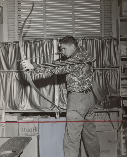 A man posing indoors with a drawn bow. He has a quiver with additional arrows affixed to his arm. Caption reads: "<b>ACCLAIMED BY ARCHERS</b> is this arm quiver, invented and produced by Robert Freid, who demonstrates its use."