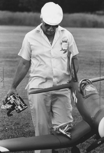 A man carrying a model airplane, possibly an F2G Corsair, and a remote control transmitter. His shirt reads: "DAVE" and has a patch depicting an airplane. Caption reads: "Dave Gauer carried his warplane off the field after if [sic] stalled and crashed during landing."