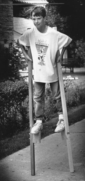 A boy walking on a pair of stilts. Caption reads: "Mark Emiley, 13, stands tall as he tries a pair of stilts during a garage sale in Cedarburg Monday."