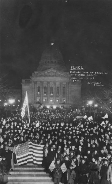 A crowd is assembled on the steps to the State Capitol. Many people are carrying American flags, and a large flag is prominently displayed. A lit "W" is at the top of the Capitol dome. Caption reads: "<u>PEACE</u> PICTURE MADE ON STEPS OF WIS-STATE CAPITOL. MON — NOV — 11th 1918 — 4 A.M. — BY — MCKILLOP & RUUD -".