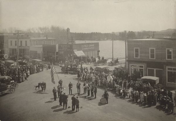 Elevated view of a flag dedication ceremony. Men holding instruments form two lines on either side of a man holding an American flag. A crowd is gathered around them. In the background is the C.I. Field Garage, and Lake Martha is behind that. Caption reads: "Lining up for the dedication of the Service Flag. Liberty day. April 26 - 18. Osseo, Wis."