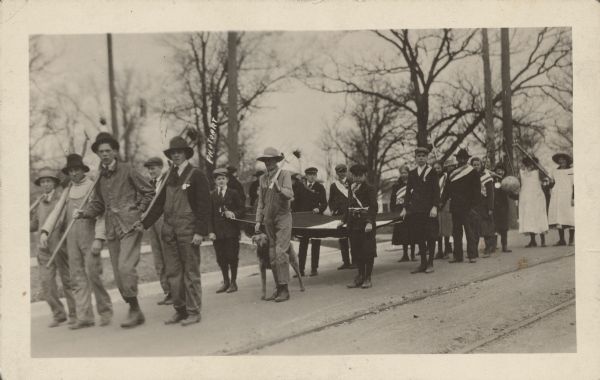 A children's parade is marching down a street. In front are several boys dressed in overalls and carrying pitchforks and other farm implements. Behind them, several boys are carrying an unidentified canvas object with a frame. Other boys and girls behind them are wearing American flag sashes. Finally, some girls are wearing white Red Cross nurse's uniform aprons. One girl is carrying a globe and appears to be wearing a fake moustache. Caption reads: "Madison, Wis. 1917. 'Hammer the Hun' parade with pupils from Randall School."
