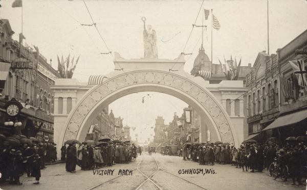 Crowds of people are standing on either side of a street, with a model of an arch spanning the street. On top of the arch is a statue of Victory. American and British flags are displayed on either side. Caption reads: "Victory Arch, Oshkosh, Wis."