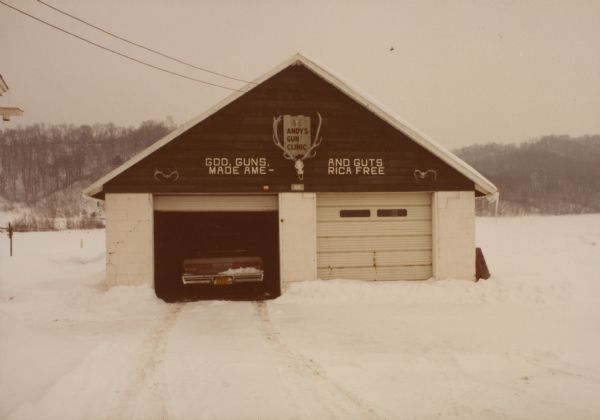 View of a two-car garage in winter with snow on the ground. One garage door is open and a car is parked inside. A sign on the garage reads: "ANDY'S GUN CLINIC" which is framed by a deer skull with antlers. Another sign above both garage doors reads: "GOD, GUNS, AND GUTS MADE AME - RICA FREE". Caption on back reads: "Feb 1983, Holland, WI. 8 mi. S. of Galesville on Hwy 53."