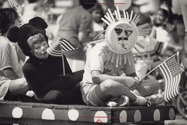 Three children are sitting on a platform. They are wearing animal costumes and holding American flags. Caption reads: "Two youngsters dressed as animals took part in the 'circus parade' portion of the parade". A headline adjacent to the image reads: "Mequon-Thiensville celebrates 4th early".