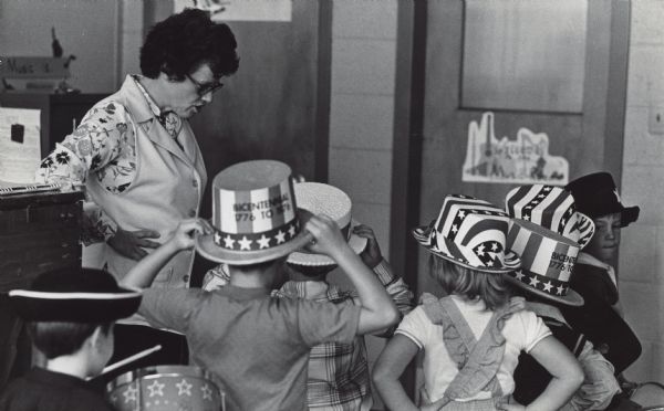 A woman is standing with her hands on her hips, looking down at a group of children. All the children are wearing hats, several of which are "Bicentennial 1776 to 1976" hats. Caption reads: "A small place for a little music. That's all that Sister Angela Esselman wanted for her class. And a music room is part of the changes in the renovated Sacred Hearts School, which has reopened in Franklin. Community News, Page 12."
