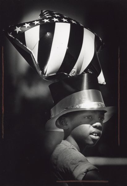 A boy is posing wearing a hat. Behind him is a star-shaped balloon with an American flag pattern. Caption reads: "Raymond Taylor, 8, waited for the judging Tuesday during a holiday bike parade in the Sherman Park neighborhood."