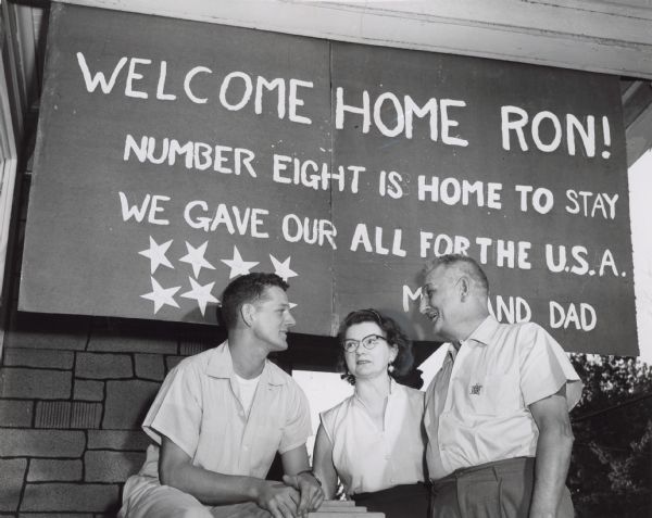 A young man is sitting next to an older man and woman. Behind them is a sign with eight stars on it, and the text:
WELCOME HOME RON! 
NUMBER EIGHT IS HOME TO STAY
WE GAVE OUR ALL FOR THE U.S.A.
MOM AND DAD

Caption reads: "<b>The last of eight sons</b> of Mr. and Mrs. Edmund Wycklendt of 2815 W. Brown st. has completed his military service. Ronald Wycklendt, 23, posed with his parents in front of a 'Welcome Home" sign at their home. Besides Ronald, who was stationed in Korea for two years, two other Wycklendt sons served in the army, two in the air force, and three in the navy. The sons all live in Milwaukee. The Wycklendts have four married daughters also."