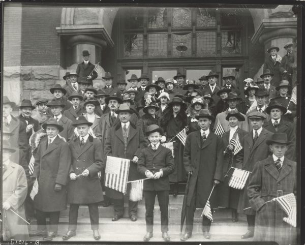 A group of people posing on the steps of a building with an stained glass arched entrance. Many of the people are holding American flags. Caption reads: "Patriotic group, otherwise "unidentified, about 1917-1918, probably in Madison Wis. ["probably" is crossed out and "Science Hall steps" has been written in.] Photo by M.E. Diemer.