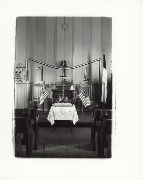 Interior view of the front of a church, with patriotic decorations. A table in the chancel has an open book surrounded by flags. On the east wall are tricolor ribbons, some of which form the numerals 76. A register of attendance and offering is in the background on the left. Caption reads: "Non Denominational Church 4 mi. west of Waupaca, WI on Hwy 54. 1976. E. Drayna, Photographer."