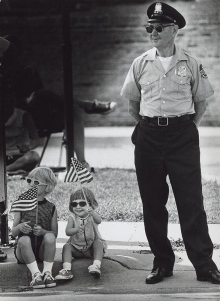 A uniformed man wearing sunglasses is standing and looking to his right. Sitting on the curb next to him are two children, who are also wearing sunglasses. The children are holding American flags. Caption reads: "George Koerner, a Wauwatosa reserve officer, found out Monday that he and sisters Stephenie and Jennifer Geyer like similar things — sunglasses and parades. They were watching the Fourth of July Parade in Wauwatosa."